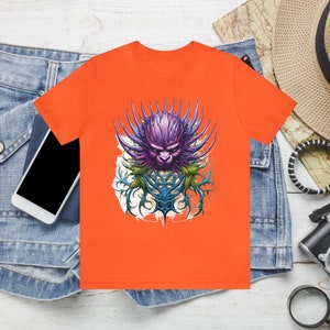 Könnte beinhalten: Orangefarbenes T-Shirt mit einem Motiv eines violetten und blauen Monsters mit Dornen und Ranken.