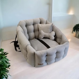 May include: A grey, quilted dog bed with a bone-shaped pillow. The bed has a black strap for securing it to a car seat.