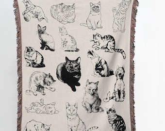 May include: A woven throw blanket with a cream background and black and white illustrations of cats in various poses. The blanket has a fringe border.