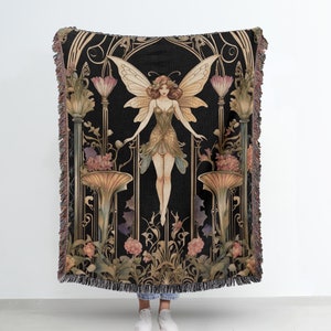 May include: A woven tapestry throw blanket features a fairy with wings and a floral design. The fairy is depicted in a green dress with a black background. The throw blanket has a fringe border.