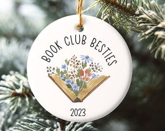 May include: White ceramic ornament with a book and flowers design. The text "Book Club Besties 2023" is printed on the ornament.