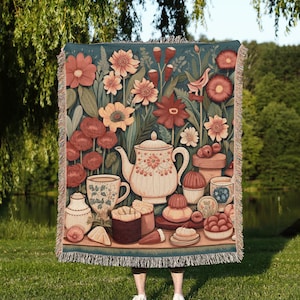 May include: A woven tapestry throw blanket with a floral design and a tea party scene. The blanket features a teapot, teacups, pastries, and flowers in shades of pink, red, and green on a teal background.