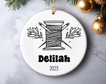 May include: White ceramic Christmas ornament with a black and white illustration of a spool of thread and two needles crossed over each other. The ornament also has the name "Delilah" and the year "2023" printed on it.