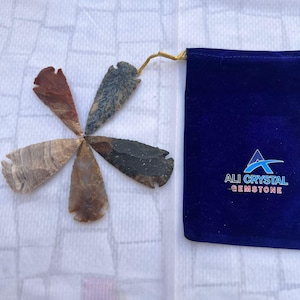 May include: A collection of six gemstone arrowheads in various colors, arranged in a circular pattern. The arrowheads are shades of brown, gray, and red. A dark blue velvet pouch with the text "ALI CRYSTAL GEMSTONE" is on the right.
