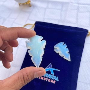 Può includere: Una mano tiene una freccia di cristallo traslucida e opalescente. Sono visibili altre due punte di freccia, una su un sacchetto di velluto blu scuro. Il sacchetto ha un logo che dice "RYSTAL GEMSTONE". I cristalli hanno un aspetto morbido e luminoso.