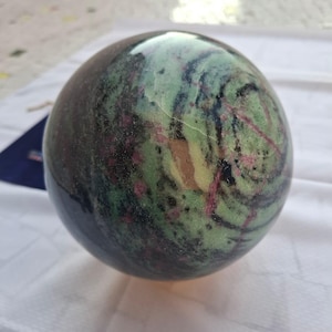 Könnte beinhalten: Eine polierte Kugel mit einer Mischung aus Farben, darunter Grün, Schwarz und Rosa. Die Kugel hat eine glatte, reflektierende Oberfläche und scheint aus einem Naturstein zu bestehen. Der Gesamteindruck ist von Tiefe und visuellem Interesse geprägt.