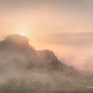 Könnte beinhalten: Landschaftsfotografie eines Berggipfels im Nebel bei Sonnenaufgang. Die Sonne ist eine helle Scheibe, die ein warmes, goldenes Licht über die Szene wirft. Das Bild ist eine Weitwinkelaufnahme, die die Weite der natürlichen Umgebung einfängt.