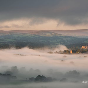 May include: A misty valley with rolling hills and a small village in the distance. The sky is a soft pink and orange, and the fog is thick and white.