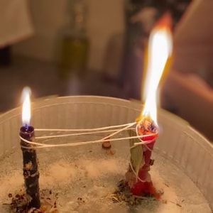 Puede incluir: Dos velas, una roja y otra negra, están ardiendo en un cuenco blanco. Las velas están atadas con un cordel blanco. El cuenco está lleno de una sustancia blanca, posiblemente arena o sal.
