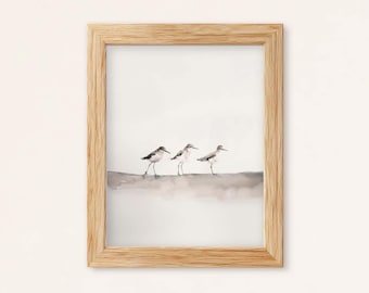 May include: A light-colored wooden frame containing a watercolor print of three small shorebirds standing in a row on a sandy beach. The birds are depicted in shades of gray and brown against a soft, neutral background with a muted wash of color.
