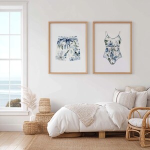 Add a touch of coastal elegance to your home with this coordinated set of two floral swimwear watercolor prints. Featuring a matching one-piece swimsuit and swim trunks adorned with soft blue florals and botanical accents, this collection blends vintage beach charm with sophisticated coastal style. Perfect for bathrooms, beach houses, lake cottages, dressing rooms, pool houses, and summer-inspired interiors.