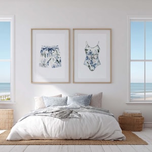 Add a touch of coastal elegance to your home with this coordinated set of two floral swimwear watercolor prints. Featuring a matching one-piece swimsuit and swim trunks adorned with soft blue florals and botanical accents, this collection blends vintage beach charm with sophisticated coastal style. Perfect for bathrooms, beach houses, lake cottages, dressing rooms, pool houses, and summer-inspired interiors.