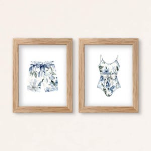 Add a touch of coastal elegance to your home with this coordinated set of two floral swimwear watercolor prints. Featuring a matching one-piece swimsuit and swim trunks adorned with soft blue florals and botanical accents, this collection blends vintage beach charm with sophisticated coastal style. Perfect for bathrooms, beach houses, lake cottages, dressing rooms, pool houses, and summer-inspired interiors.