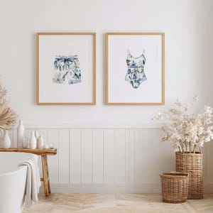Add a touch of coastal elegance to your home with this coordinated set of two floral swimwear watercolor prints. Featuring a matching one-piece swimsuit and swim trunks adorned with soft blue florals and botanical accents, this collection blends vintage beach charm with sophisticated coastal style. Perfect for bathrooms, beach houses, lake cottages, dressing rooms, pool houses, and summer-inspired interiors.