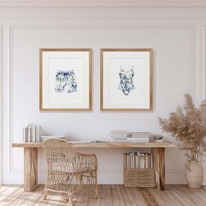 Add a touch of coastal elegance to your home with this coordinated set of two floral swimwear watercolor prints. Featuring a matching one-piece swimsuit and swim trunks adorned with soft blue florals and botanical accents, this collection blends vintage beach charm with sophisticated coastal style. Perfect for bathrooms, beach houses, lake cottages, dressing rooms, pool houses, and summer-inspired interiors.