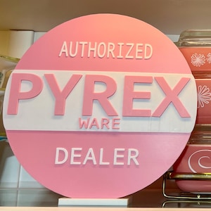 May include: A pink round sign with white lettering that reads "AUTHORIZED PYREX WARE DEALER".  Three pink Pyrex baking dishes with white floral designs are stacked on a gold metal stand.