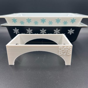May include: White plastic baking dish stand with the numbers 548 and 575 printed on the side. The stand is designed to hold two white and black baking dishes with a snowflake pattern.