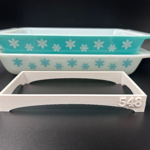 May include: Two turquoise blue glass baking dishes with a white snowflake pattern. The dishes are stacked on top of each other. A white plastic baking dish rack with the number 548 printed on the side is in front of the dishes.
