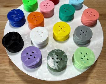 May include: A collection of fifteen cylindrical, button-shaped objects in various colors, including red, blue, green, orange, pink, yellow, white, black, purple, and gray. Each object features a ribbed side and a flat top with five small holes.