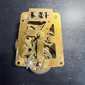 May include: A brass clock movement mechanism featuring an intricate arrangement of interlocking gears, wheels, and a central arbor plate. The metallic structure displays a weathered patina with visible screws and various sized cogs.