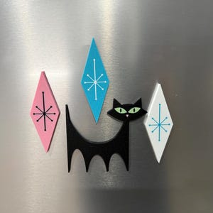 May include: A set of four decorative magnets featuring a black cat silhouette with green eyes, accompanied by three diamond-shaped pieces in pink, blue, and white, each decorated with a thin black or blue starburst pattern.