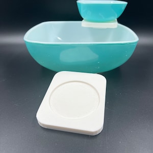 May include: A turquoise blue ceramic serving dish with a smaller turquoise blue bowl on top. The dish has a square shape and the bowl is round. There is a white ceramic base for the bowl to sit on.