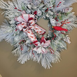 Op de afbeelding: Een bevroren kerstkrans met een grote strik met rode kardinalen. De krans is versierd met rode bessen, dennenappels en een rood kardinaalbeeldje. De basis van de krans is gemaakt van bevroren dennennaalden.