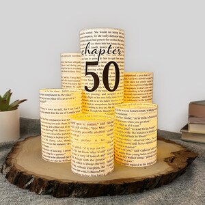 May include: A set of five cylindrical paper luminaries featuring printed vintage book text. The tallest central cylinder displays the black text "chapter 50" in a decorative font. The paper shades are arranged in a tiered formation on a wood slice.