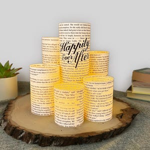 May include: A set of five cylindrical luminaries made from vintage book pages, featuring the text "Happily Ever After" in a black script font on the tallest piece. Each light-colored paper shade displays printed literary text.