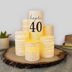 May include: A set of five cylindrical luminaries crafted from vintage book pages, featuring a central taller piece with the black text "chapter 40" printed on the front. The paper shades emit a warm, soft glow, showcasing printed literary text.