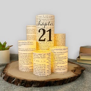 May include: A set of five cylindrical paper luminaries featuring printed book text and a large black number 21 on the central piece. The cream-colored paper wraps display various paragraphs of English literature text in a dark serif font.