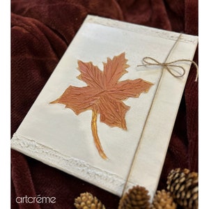 Puede incluir: Un lienzo blanco con un borde texturizado, que presenta una sola hoja de arce marrón anaranjado con un tallo dorado. La hoja está pintada en un estilo en relieve texturizado, dándole una apariencia tridimensional. Un lazo de cuerda marrón está atado alrededor del lienzo.