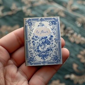 May include: A small, blue and white patterned book with a gold frame and the text "la fleur" on the front cover.