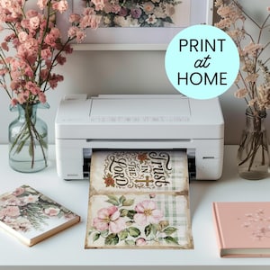 May include: A white printer is printing two floral-themed cards. The top card reads "Trust in the Lord" in a decorative font. The bottom card features pink flowers. The image also includes a light blue circle with the words "PRINT at HOME".
