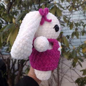 May include: White crocheted bunny wearing a pink dress and a pink bow. The bunny has black eyes and a white fluffy tail.