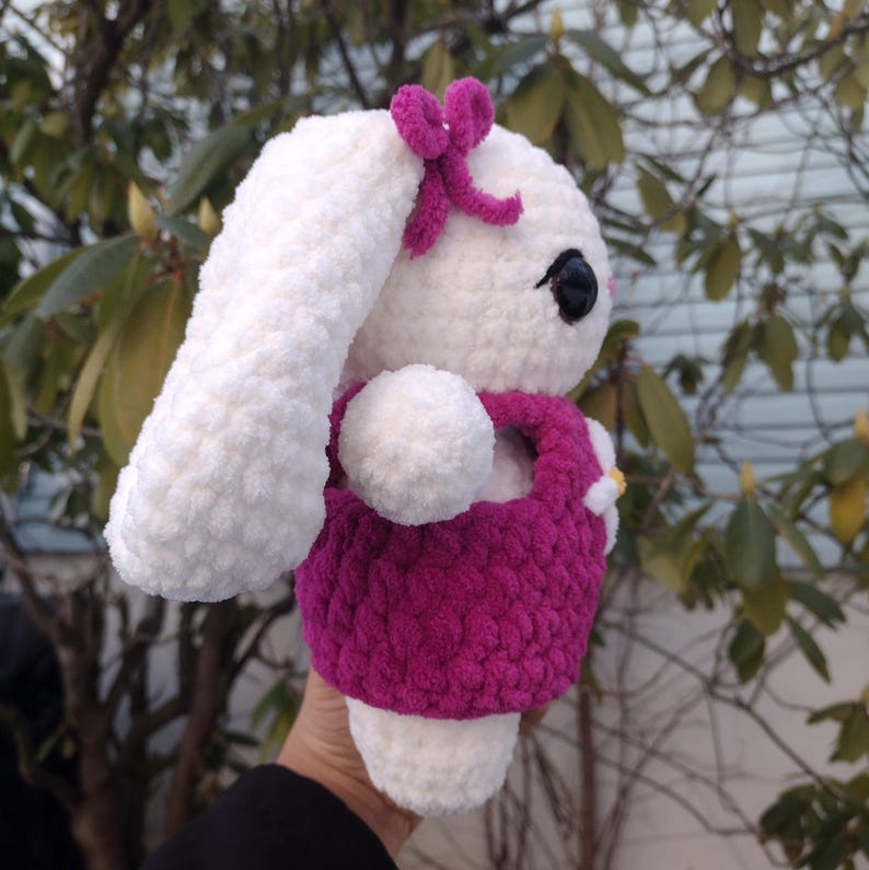 May include: White crocheted bunny wearing a pink dress and a pink bow. The bunny has black eyes and a white fluffy tail.