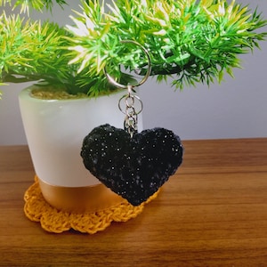May include: A black crocheted heart keychain with a silver chain attached to a key ring. The keychain is hanging from a small potted plant.