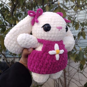 May include: White crocheted bunny plush toy wearing a pink dress with a white flower and yellow centre. The bunny has black eyes and a pink nose.