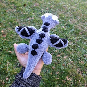 May include: A crocheted plush dragon toy with gray fur and black wings. The dragon has a white snout and black eyes.