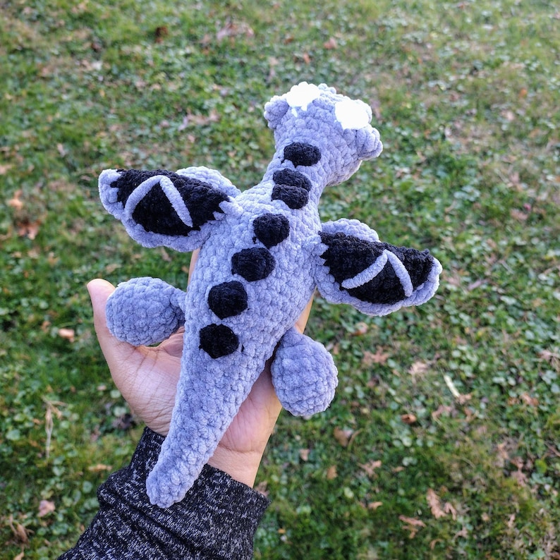 May include: A crocheted plush dragon toy with gray fur and black wings. The dragon has a white snout and black eyes.