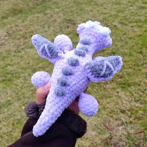 May include: A crocheted purple dragon toy with gray wings and a white fluffy head. The dragon is held in a person's hand.