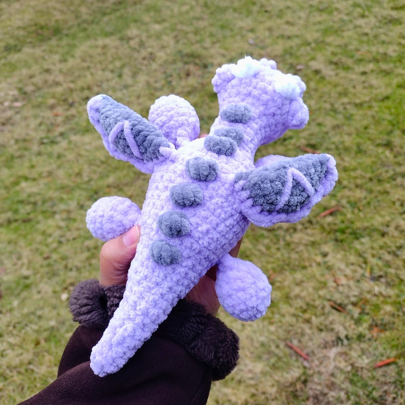 May include: A crocheted purple dragon toy with gray wings and a white fluffy head. The dragon is held in a person's hand.