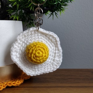 May include: A white crocheted fried egg keychain with a yellow yolk. The keychain is attached to a silver key ring.