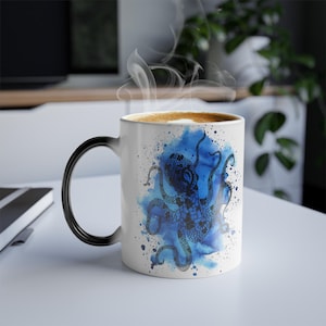 Puede incluir: Taza de cerámica blanca con asa negra, con un diseño de pulpo en acuarela azul. El pulpo está centrado en una salpicadura azul con detalles de salpicaduras de tinta. La taza contiene una bebida caliente con espuma.