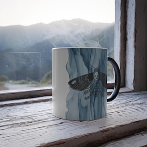 Puede incluir: Una taza de cerámica blanca con asa negra presenta una ilustración detallada de una polilla con diseño de calavera. La polilla está sobre un fondo azul acuarela. Sale vapor de la taza.