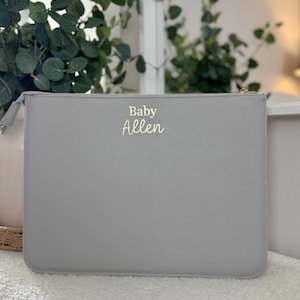 May include: A pale grey clutch bag with a gold zip and the name "Baby Allen" in gold script. The rectangular bag is made of a smooth material. Greenery and a white surface are visible in the background.