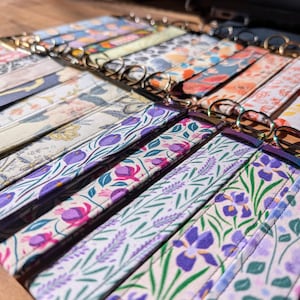 May include: A selection of fabric wrist lanyards with floral designs. The lanyards display patterns of purple irises, lavender, and other colorful flowers. Each lanyard is fitted with a gold-coloured metal ring.