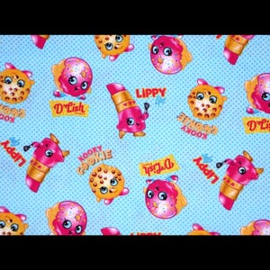 May include: Light blue fabric with a repeating pattern of cartoon characters. The characters include donuts, lipstick, and cookies, with the words "D'Lish," "Kooky," and "Lippy" on the fabric. The background has a small polka dot pattern.