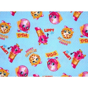 May include: A light blue fabric with a repeating pattern of cartoon characters. The characters include donuts, cookies, and lipstick tubes, each with smiling faces and bright colors. Text includes "D'Lish Donut", "Kooky Kookie", and "Lippy Lips".