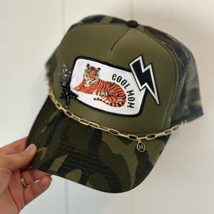 May include: Camouflage trucker hat with a tiger patch that reads "COOL MOM" and a lightning bolt. The hat has a gold chain detail and a black star embellishment. The hat is olive green and brown.